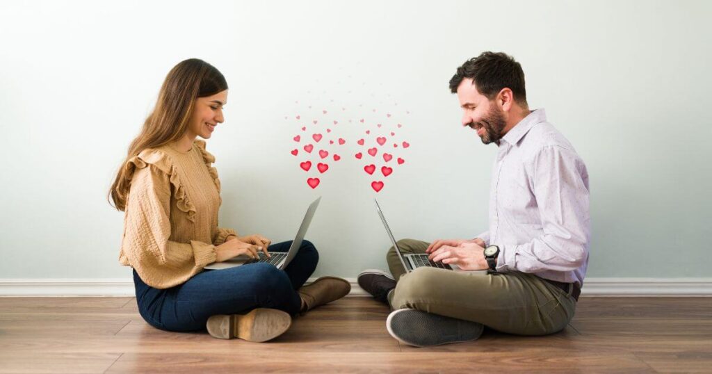 An image of a man and woman sitting on the ground cross-legged and working on their laptops, facing one another. They are looking at the laptops and smiling, and graphics of red hearts are coming out from each laptop.