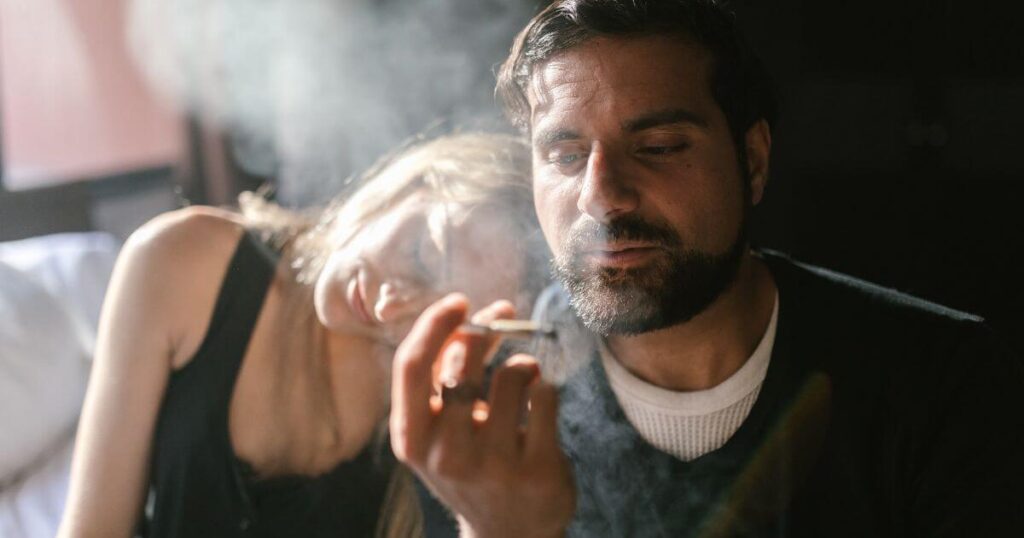 A man and a woman pensively sitting on a bed while smoking cannabis.