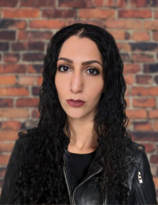 A picture of Lilithfoxx wearing a black leather jacket and standing in front of a red brick wall.