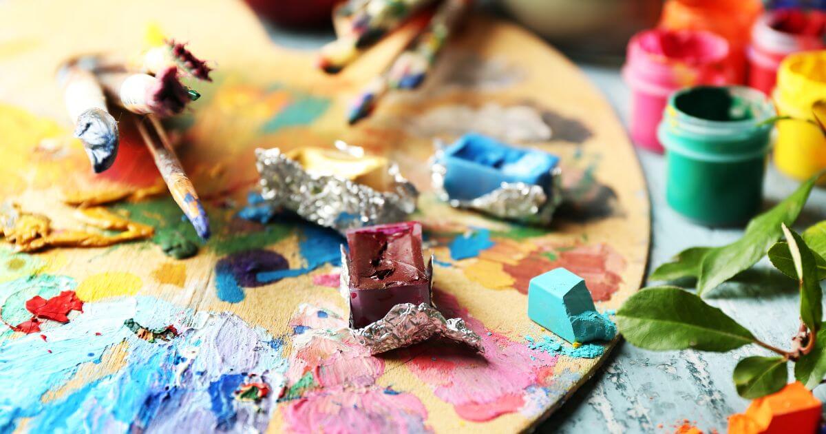 An image of an artist's paint palette with multiple paints, colors, brushes, and mediums scattered on it.