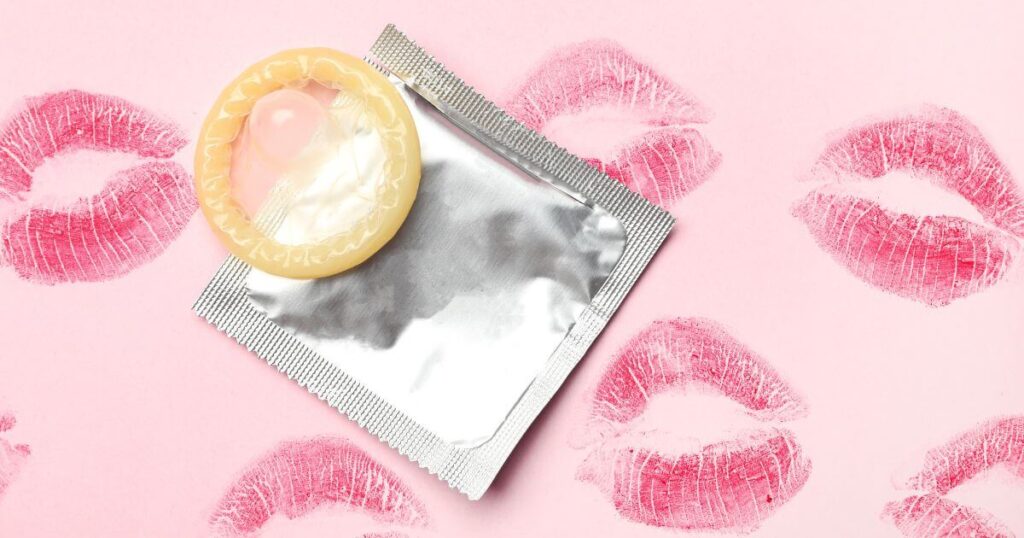 A pink background with red lipstick kisses all over it. Sitting on the background there is an unused external condom and its package.