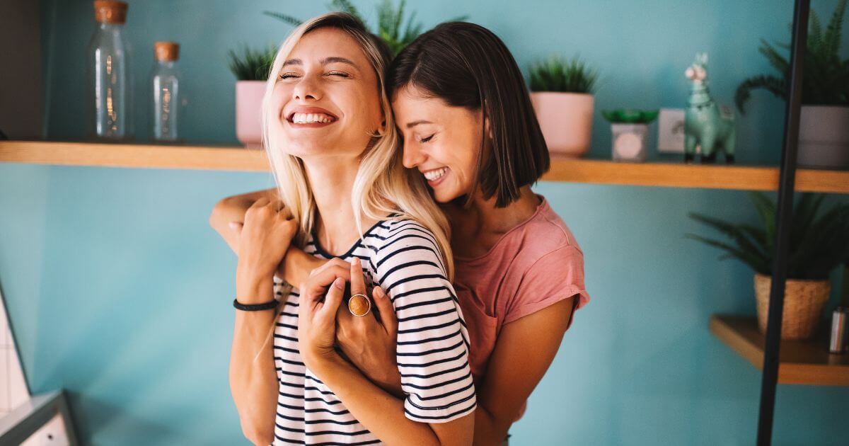 A lesbian couple enjoying the benefits of effective healthy communication in their relationship. They are in an embrace, smiling, and standing in their kitchen on a bright sunny day.