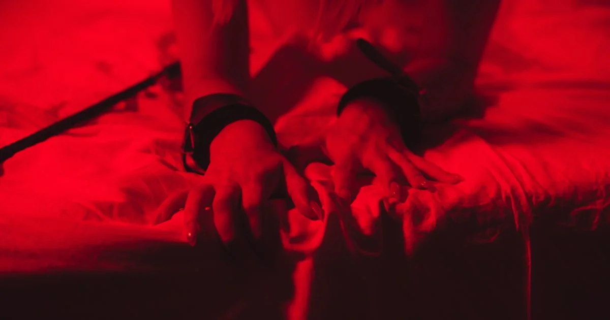 Hands restrained with cuffs in red lighting during a BDSM scene, representing a first BDSM experience and consensual kink exploration.