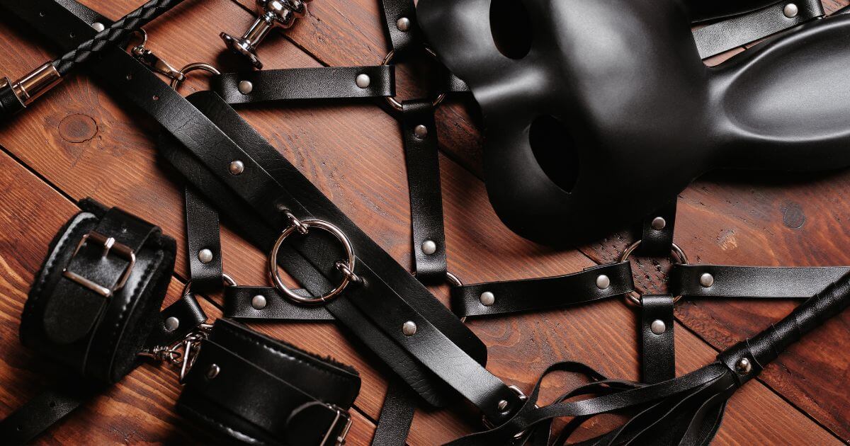 What does BDSM stand for? Black leather BDSM restraints, cuffs, harnesses, and masks arranged on a wooden table.